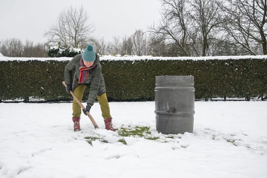 a middle-aged woman is collecting snow in a barrel with a shovel. For further watering plants in a greenhouse, the concept of protecting the environment and conserving natural resources