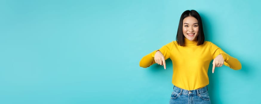 Shopping concept. Beautiful korean girl with happy smile, pointing fingers down at banner, standing against blue background.