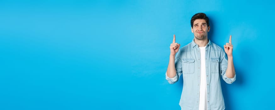 Portrait of doubtful adult man looking disappointed, pointing fingers up at something with uncertain face, standing over blue background.