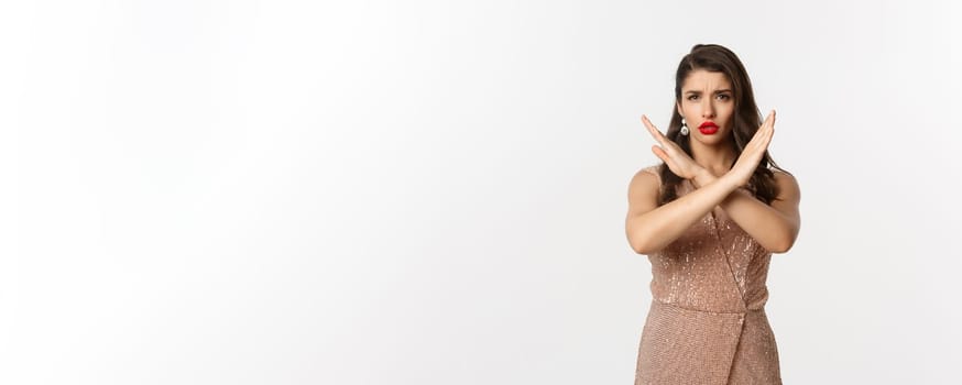 Displeased young woman in luxurious dress and red lipstick saying no, showing stop sign, cross gesture to refuse or prohibit, standing over white background.