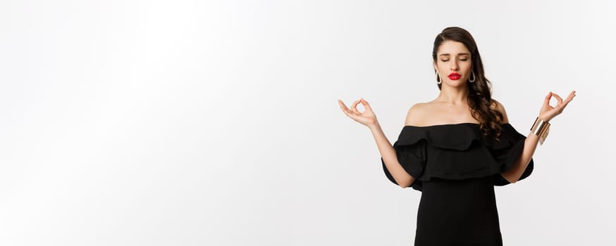 Calm and patient young brunette female meditating, standing in black dress with hands in zen gesture, standing against white background.