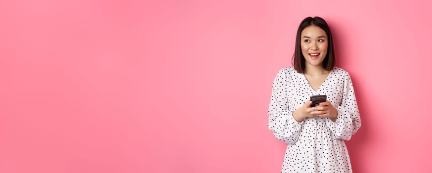 Cute asian woman thinking and smiling, looking left dreamy while messaging on smartphone, browing online stores, standing over pink background.