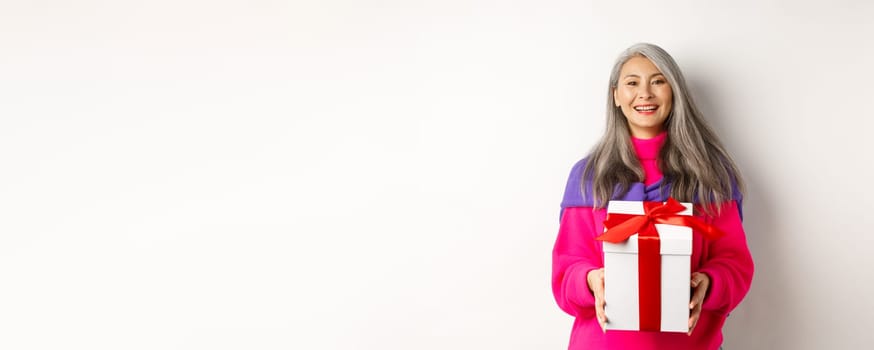 Beautiful asian senior woman smiling, congratulating with valentines day, holding gift in box, standing over white background.