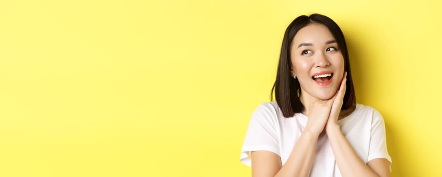 Cute romantic girl in white t-shirt imaging valentines day, looking left with admiration and joy, smiling with hands pressed to cheek, standing over yellow background.