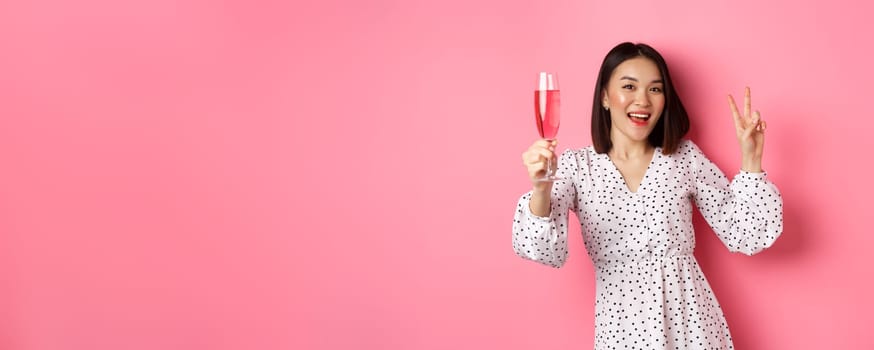 Cute asian female model drinking champagne, celebrating on party and showing peace sign, smiling happy at camera, standing over pink background.