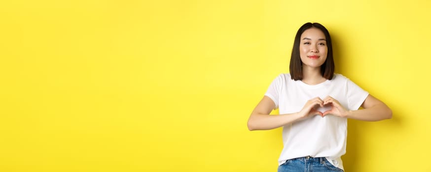 Beautiful asian woman showing I love you heart gesture, smiling at camera, standing against yellow background. Concept of valentines day and romance.