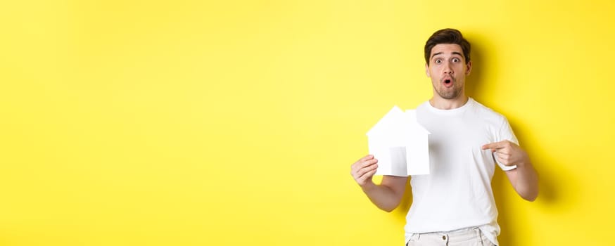 Real estate concept. Excited man pointing finger at paper house model, searching for flat, standing over yellow background.