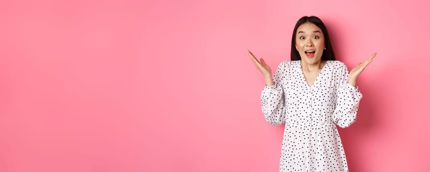 Happy and surprised asian woman rejoicing, spread hands and gasping amazed, looking with excitement and disbelief, standing over pink background.