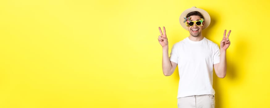 Concept of tourism and lifestyle. Happy man enjoying trip, wearing summer hat and sunglasses, posing with peace signs for photo, yellow background.