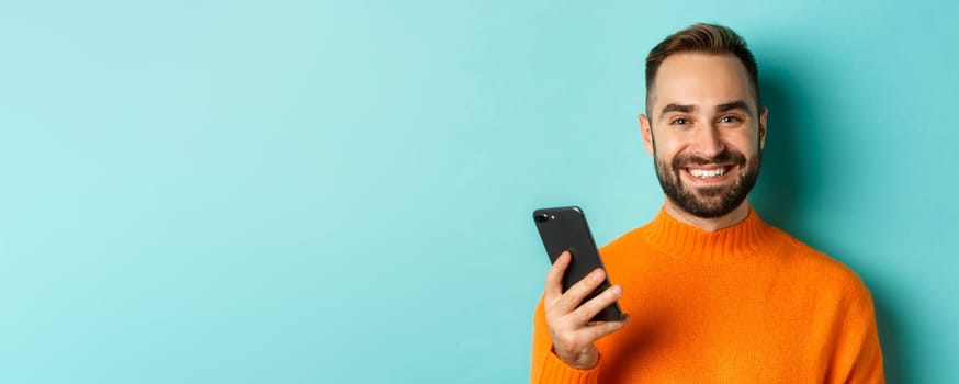 Close-up of happy handsome man writing message on mobile phone, holding smartphone and smiling, standing against turquoise background.