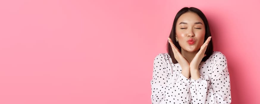 Beauty and skin care concept. Close-up of beautiful asian girl close eyes, pucker lips for kisses, showing cute perfect face, standing over pink background.