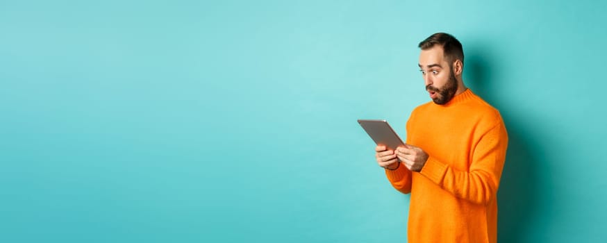 Image of male model in orange sweater staring at digital tablet screen, looking surprised, standing over light blue background.