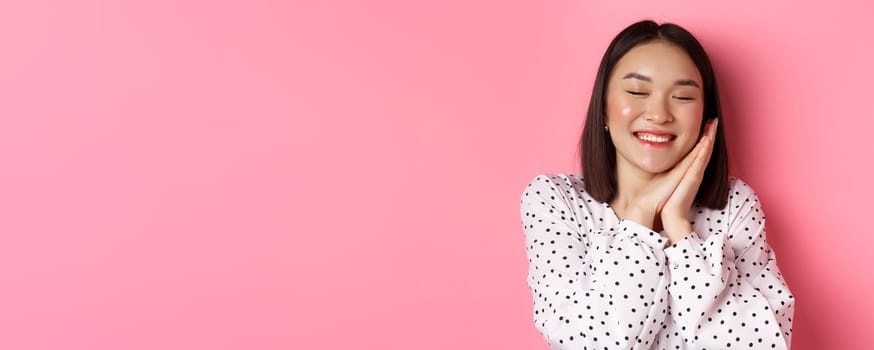 Beauty and lifestyle concept. Close-up of beautiful and dreamy asian woman sleeping on her hands, close eyes and smiling, daydreaming on pink background.