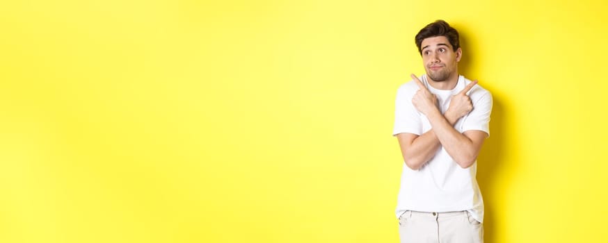 Indecisive man shrugging, pointing fingers sideways, troubled to choose, standing over yellow background.