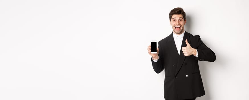 Image of handsome male entrepreneur in black suit, recommending app or online shop, showing thumbs-up and smartphone screen, standing over white background.