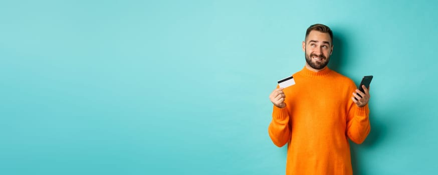 Online shopping. Thoughtful man holding credit card and mobile phone, thinking of purchase, standing over light blue background.