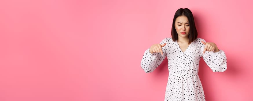 Concerned asian woman peeking down and pointing hand at disturbing banner, frowning upset, standing over pink background.