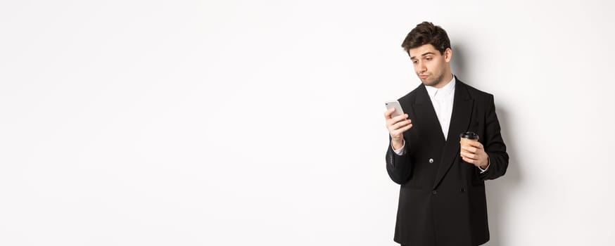 Portrait of thoughtful handsome businessman, drinking coffee and browsing in internet, looking at smartphone screen, standing against white background.