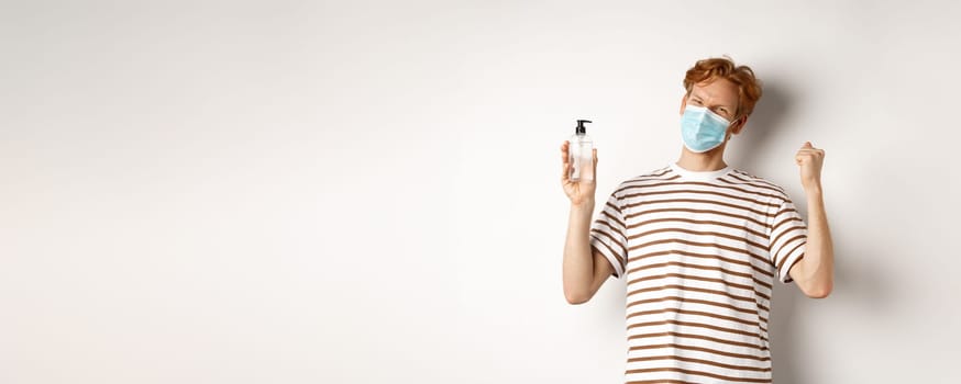 Covid-19, health and lifestyle concept. Handsome young man with red hair wear face mask and celebrating, showing hand sanitizer and looking cheerful, white background.