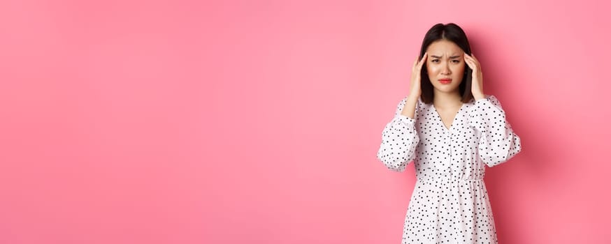 Image of upset asian woman having headache, feeling unwell or dizzy, close eyes and massaging head, suffering migraine, standing over pink background.