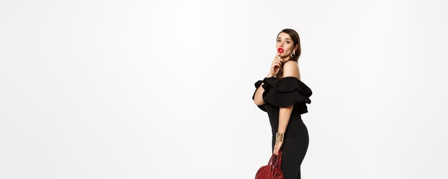 Beauty and fashion concept. Full length of sensual young woman in black elegant dress, high heels and purse, touching lip flirty and gazing at camera, white background.