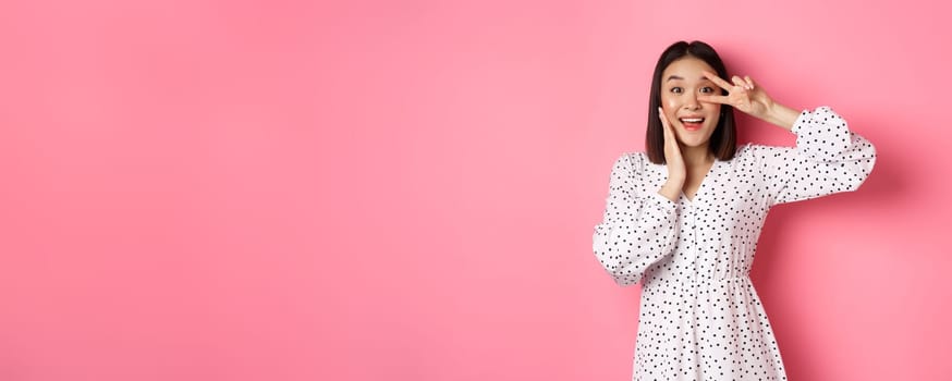 Beautiful and feminine asian woman showing kawaii sign on eye and looking amazed at camera, standing in dress over pink background.