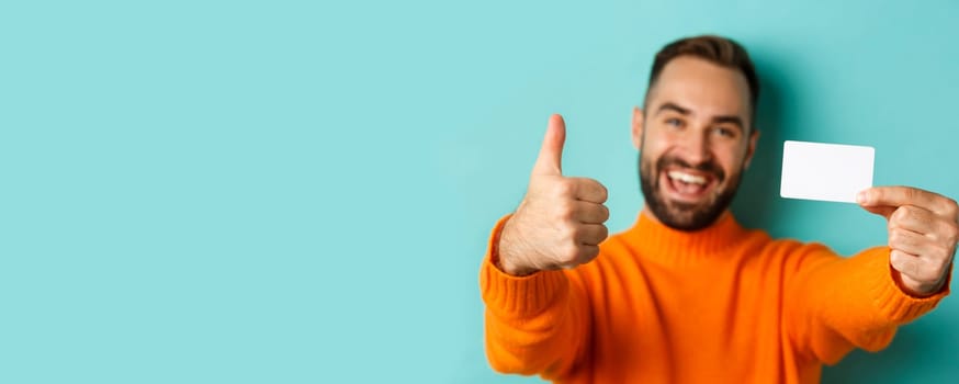 Close-up of handsome caucasian man going on shopping, showing credit card and thumb up in approval, standing over turquoise background.
