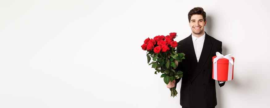 Concept of holidays, relationship and celebration. Handsome boyfriend in black suit, holding bouquet of red roses and a gift, wishing merry christmas, standing over white background.