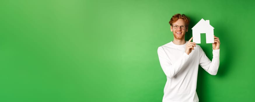 Real estate. Handsome young man with red hair, wearing glasses, showing paper house cutout and smiling, standing against green background.