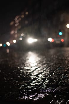 Blurred low angle view of car headlights shining in rain with reflection on wet cobbled at night.