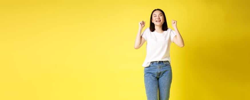 Full size shot of cheerful asian woman in jeans and white t-shirt jumping from happiness, winning prize and rejoicing, celebrating and shouting yes with joy, yellow background.