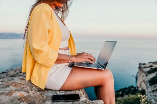 Successful business woman in yellow hat working on laptop by the sea. Pretty lady typing on computer at summer day outdoors. Freelance, travel and holidays concept.