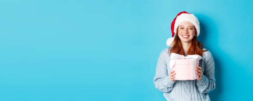 Winter holidays and Christmas Eve concept. Smiling redhead girl in sweater and Santa hat, holding New Year gift and looking at camera, standing against blue background.