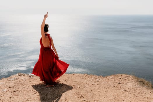 Side view a Young beautiful sensual woman in a red long dress posing on a rock high above the sea during sunrise. Girl on the nature on blue sky background. Fashion photo.