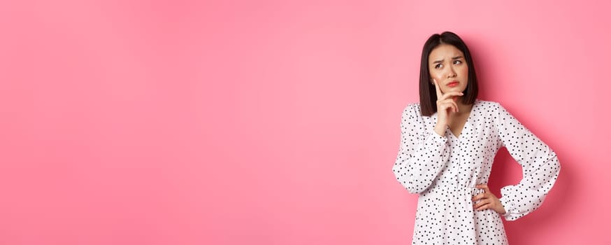 Concerned young asian female model making decision, seriously thinking, looking at upper left corner and choosing, standing over pink background.