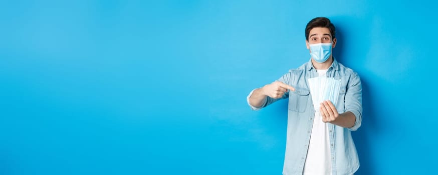 Concept of coronavirus, quarantine and social distancing. Young man pointing at medical masks, preventing measures from covid-19, standing over blue background.