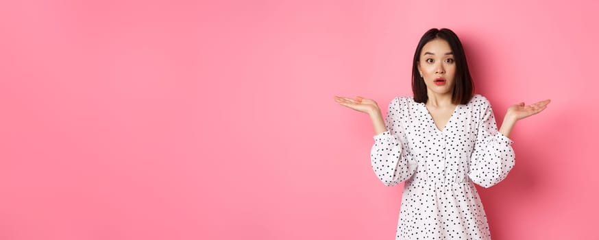 Confused and clueless asian girl shrugging, staring at camera puzzled, dont know what to do, spread hands sideways, standing over pink background.