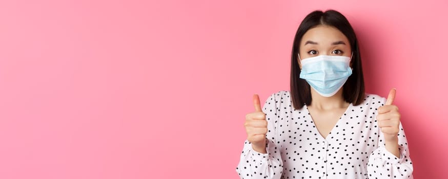 Covid-19, pandemic and lifestyle concept. Supportive asian female in face mask showing thumbs-up, approve and praise good work, standing over pink background.