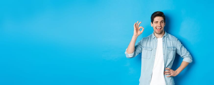 Confident adult man winking and showing okay sign, approve something good, standing against blue background.