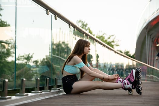 A long-haired girl sits on a bridge and puts on rollerskates. She has a glass railing behind her back, in which the reflection of the girl can be seen. The woman is wearing a blue T-shirt and black shorts.
