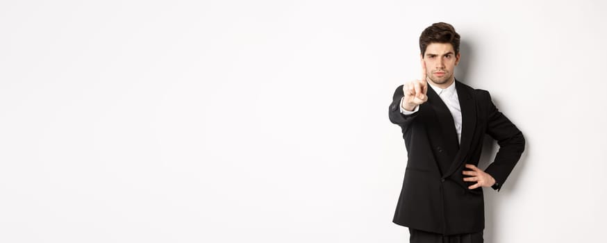 Portrait of serious handsome man in business suit, showing one finger to prohibit or decline something, telling to stop, disagree with you, standing over white background.