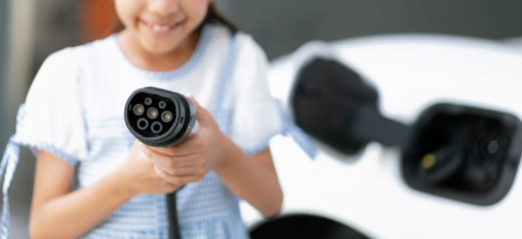 Focus electric charger plug from home charging station for EV engine power by renewable energy with blur background of girl pointing charger device at camera. Concept of progressive young generation.