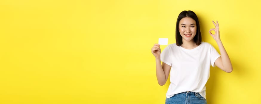 Young asian woman in casual white t-shirt showing plastic credit card and okay gesture, recommend bank, smiling at camera, yellow background.