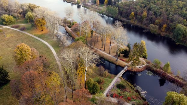 Flying over autumn park. Many trees with yellow green and fallen leaves, lakes, river, people walking along dirt paths in park on autumn day. Top view. Aerial drone view. Beautiful natural background