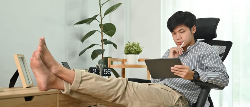 Casual man freelancer sitting on comfortable chair in modern home office and using digital tablet.