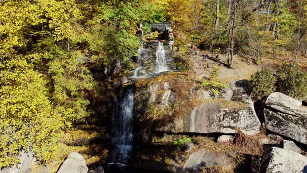 Waterfall in autumn park. Water falls from high stones, trees with yellow leaves in landscape park on sunny autumn day. Fallen leaves, flowing water, forest. Natural landscape, nature scenery. Aerial