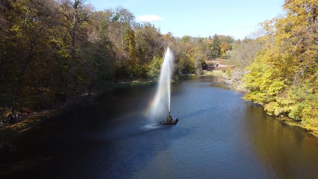 Decorative fountain with high jet of water in center of lake among trees with yellow leaves in landscape park on sunny autumn day. Falling drops of fountain create rainbow. People walking park paths