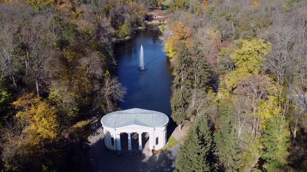 Beautiful landscaped park with lake, fountain, trees and white architecture on sunny autumn day. Aerial drone view. Trees with yellow green leaves on shores and high flowing jet of fountain.