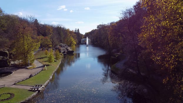 Beautiful landscaped park with lake, fountain, trees, large stones and lot of people walking on sunny autumn day. Trees yellow leaves on shores, tourists, flowing jet of fountain.