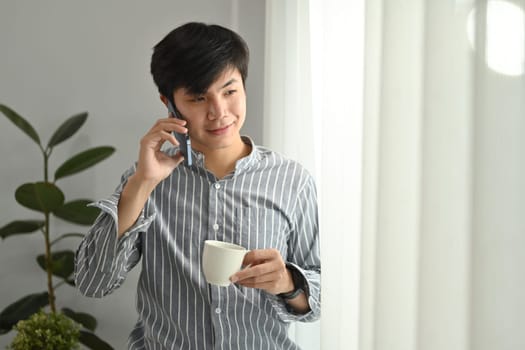 Portrait of smiling asian businessman holding coffee cup and talking on mobile phone at bright home office.